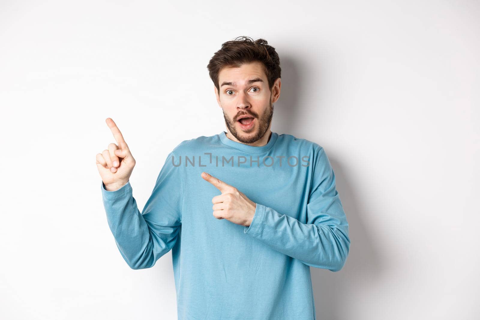 Amazed man showing awesome promo offer, pointing fingers at upper left corner logo and looking excited, recommend product, white background by Benzoix