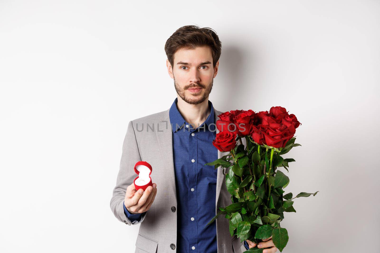 Handsome man in suit going to make a proposal, standing with red roses flowers and engagement rin in box, making romantic surprise, white background.