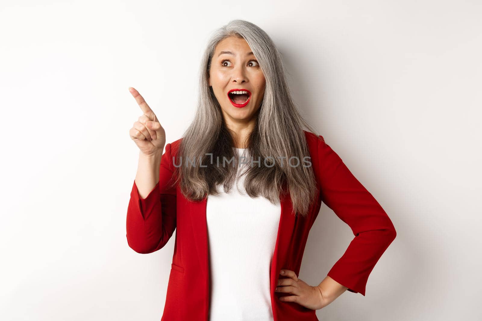 Surprised senior asian lady pointing upper left corner, checking out promo offer and smiling amazed, standing in red blazer over white background by Benzoix