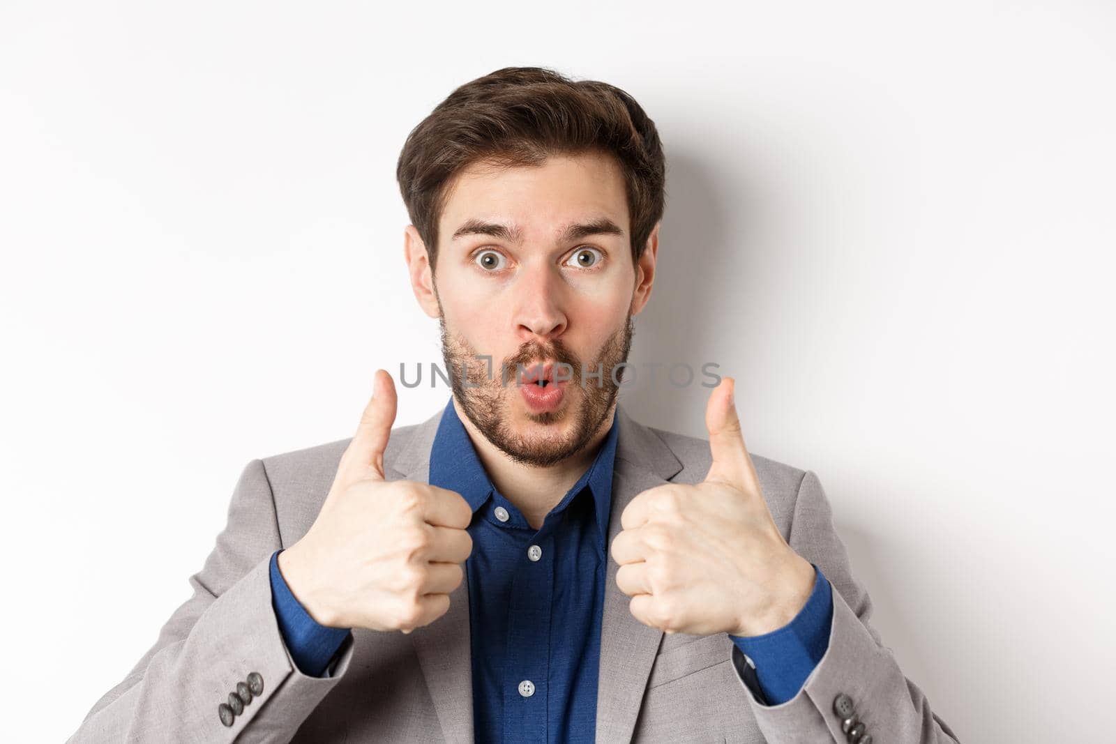 Close up portrait of excited guy in suit say wow, showing thumbs up with amazed face, checking out awesome promo, white background.