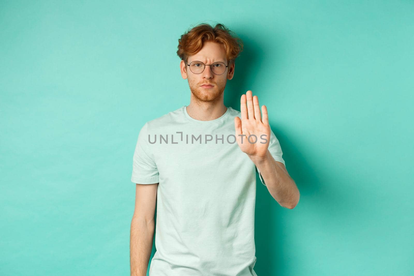 Angry and serious young man with red hair, wearing glasses, showing stop gesture, telling no, disapprove and prohibit something bad, standing over turquoise background.