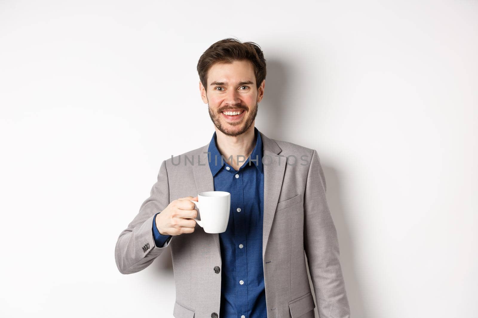 Handsome businessman in suit drinking coffee or tea from office mug, smiling enthusiastic at camera, standing against white background.