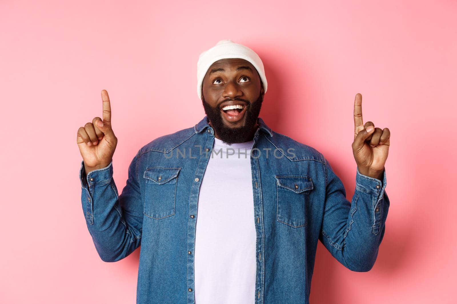 Happy african american man showing advertisement, smiling amazed and staring at camera, pointing fingers up, pink background.