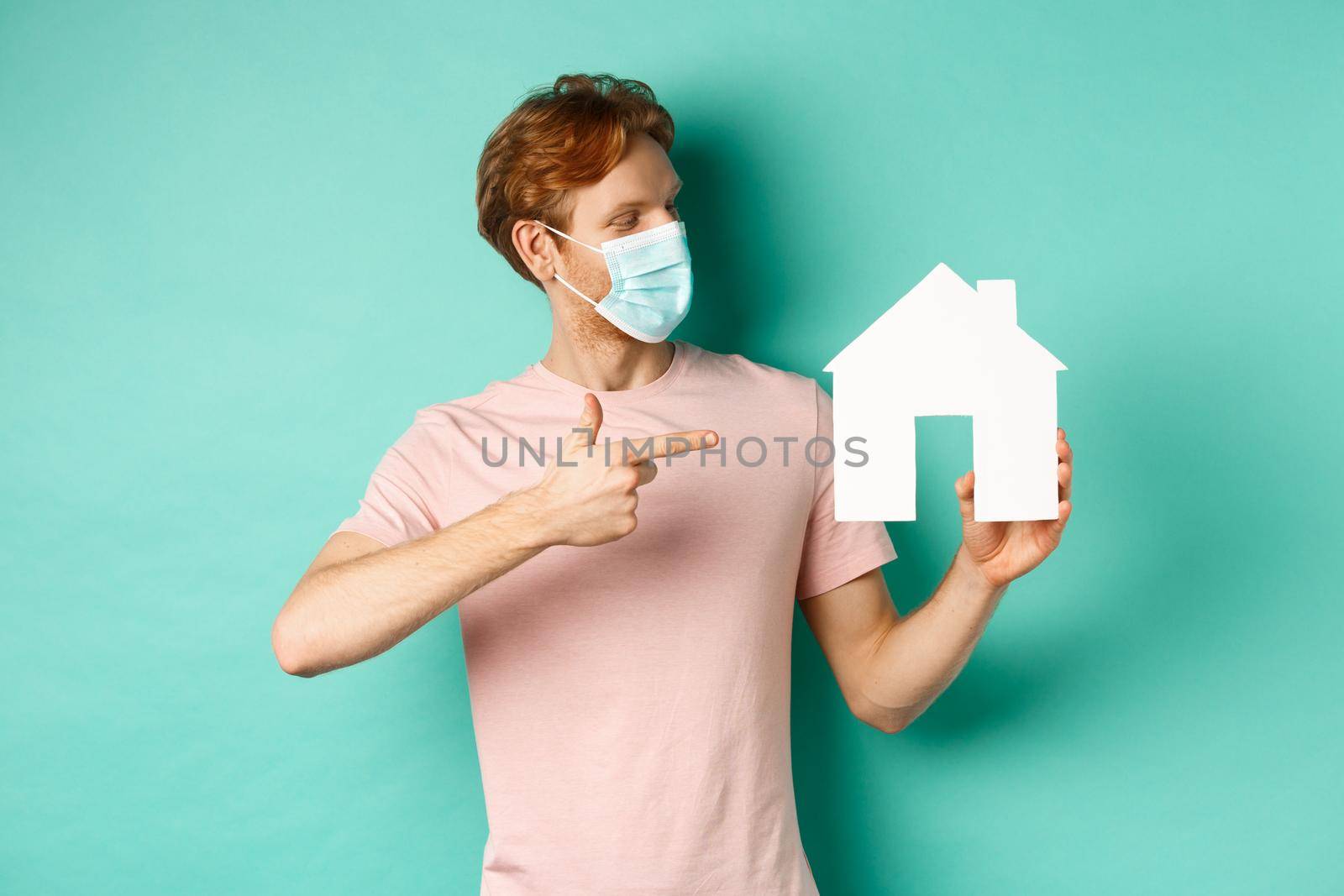 Covid-19 and real estate concept. Redhead guy in medical mask pointing and looking at paper house cutout, standing over turquoise background.