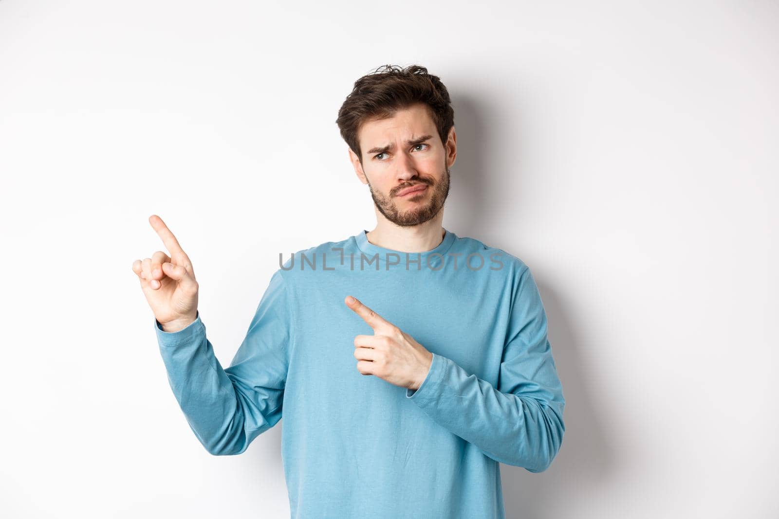 Skeptical and unimpressed young man grimacing, pointing fingers and looking at bad promotion, standing dissatisfied over white background by Benzoix