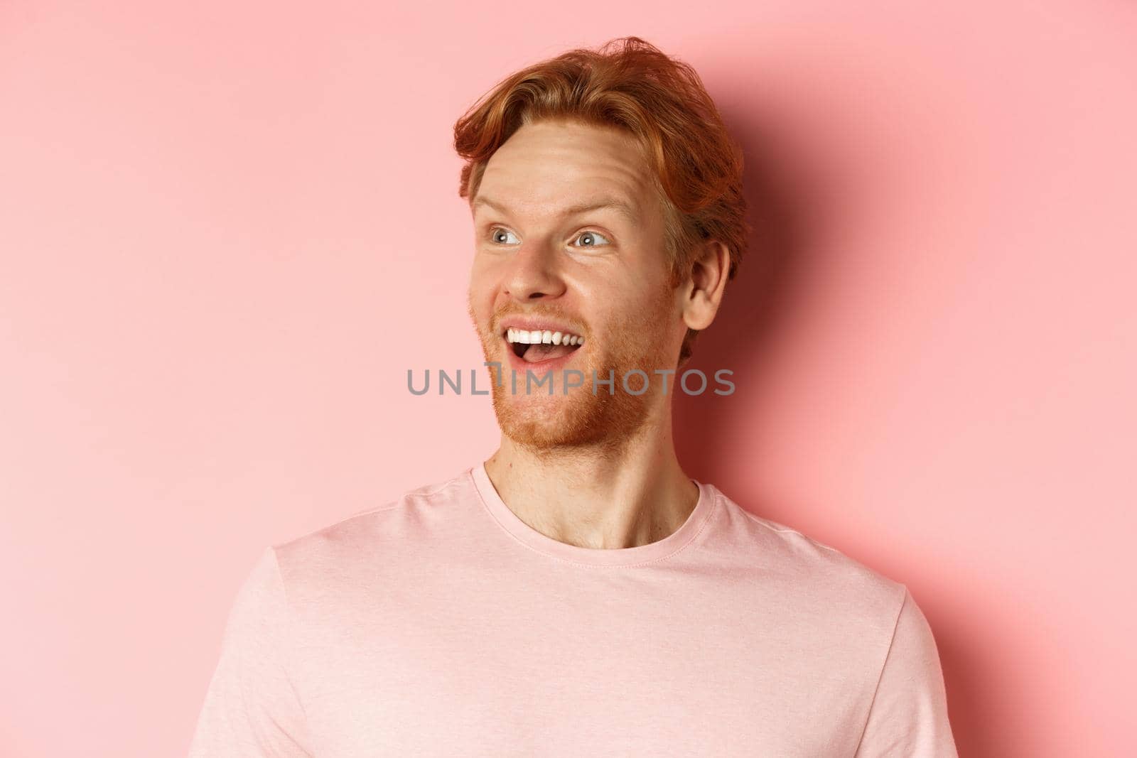 Close-up of handsome caucasian guy with red hair and beard, looking left with happy smile, checking out promo offer, standing over pink background by Benzoix