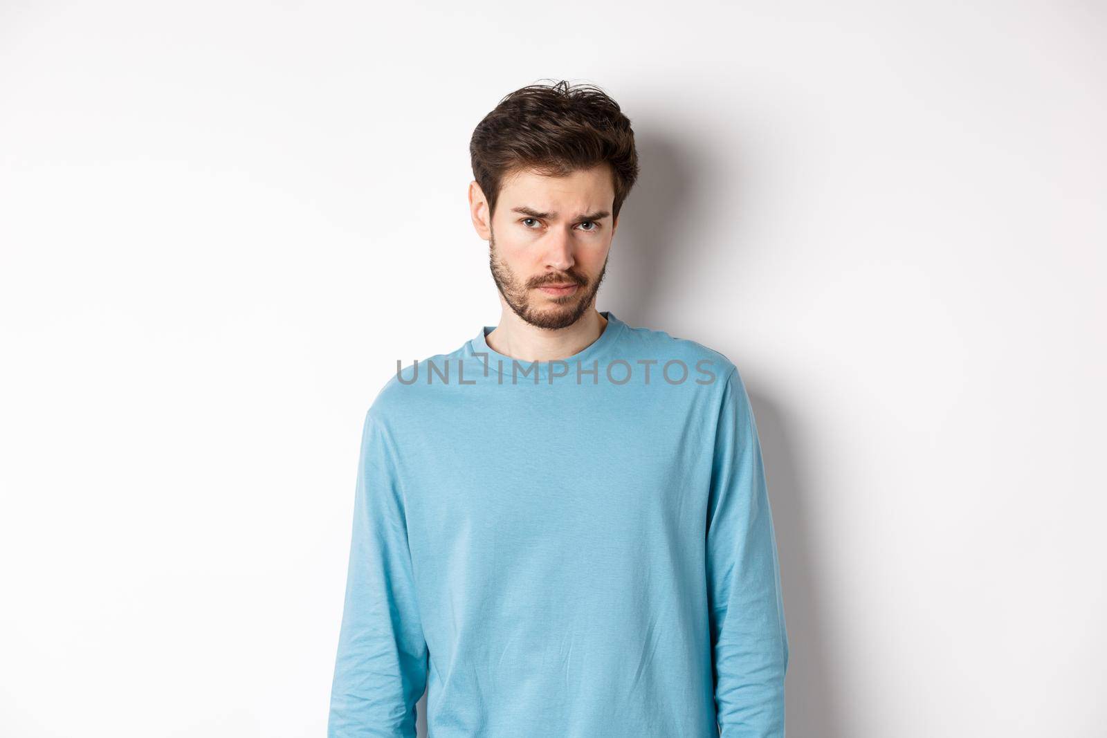 Sad young bearded man sulking, looking upset at camera, offended by someone, standing against white background by Benzoix