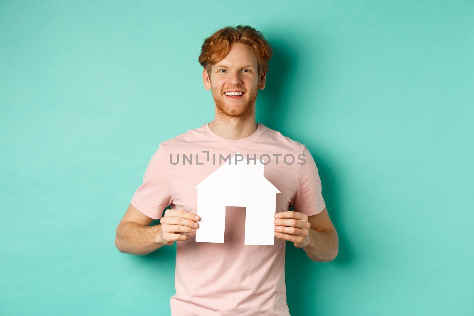 Real estate concept. Young man with red hair, wearing t-shirt, showing paper house cutout and smiling happy, standing over mint background. Copy space