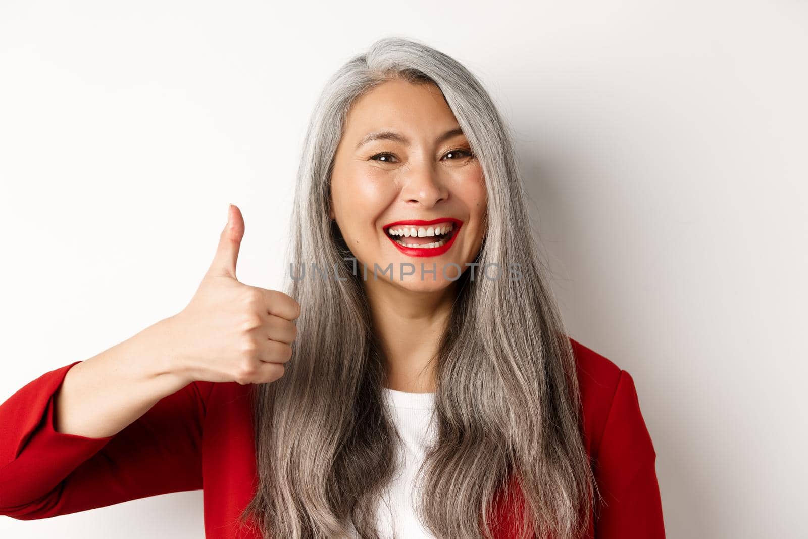 Close-up of happy asian female manager in red blazer, showing thumb-up and smiling in approval, like and agree, white background.