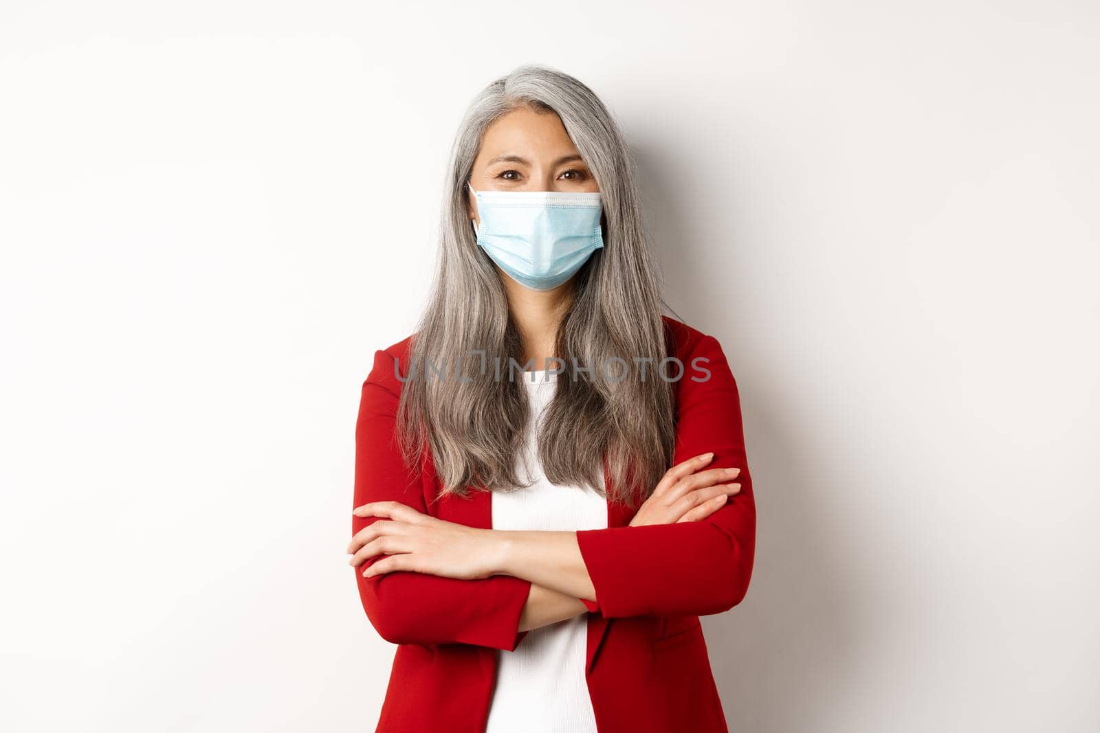 Covid-19 and business people concept. Cheerful asian lady in face mask looking at camera, cross arms on chest like professional, white background.