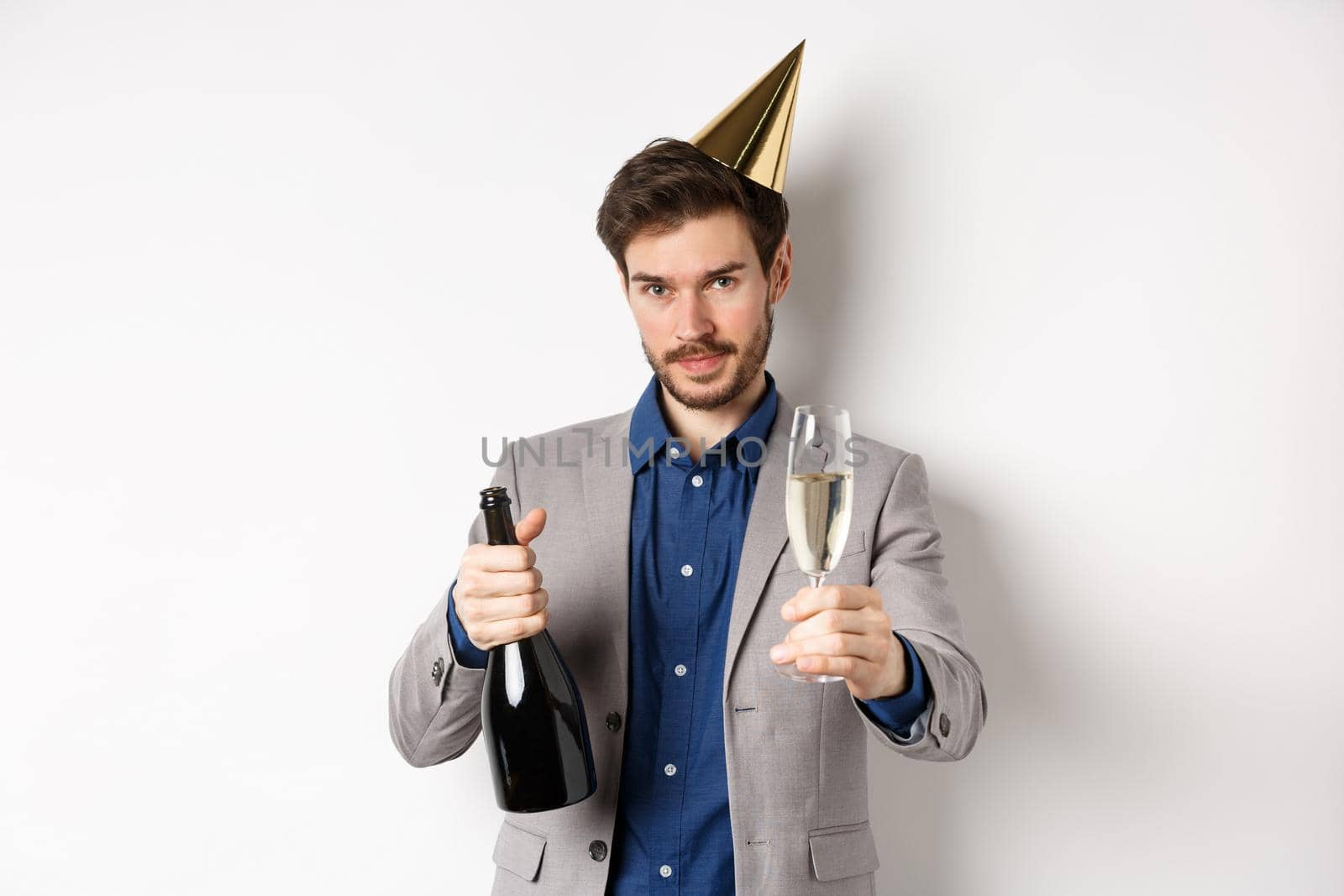 Celebration and holidays concept. Handsome man in suit and birthday hat giving glass of champagne, holding bottle alcohol, standing on white background.
