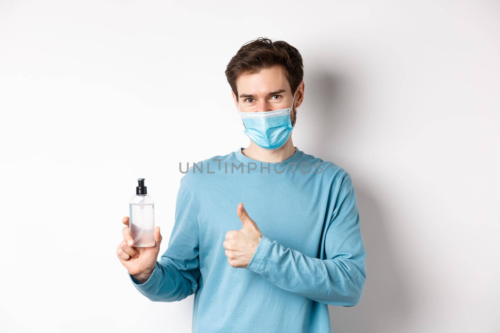 Covid-19, health and quarantine concept. Handsome smiling man in face mask, showing thumbs up and hand sanitizer bottle, recommending antiseptic brand, white background.