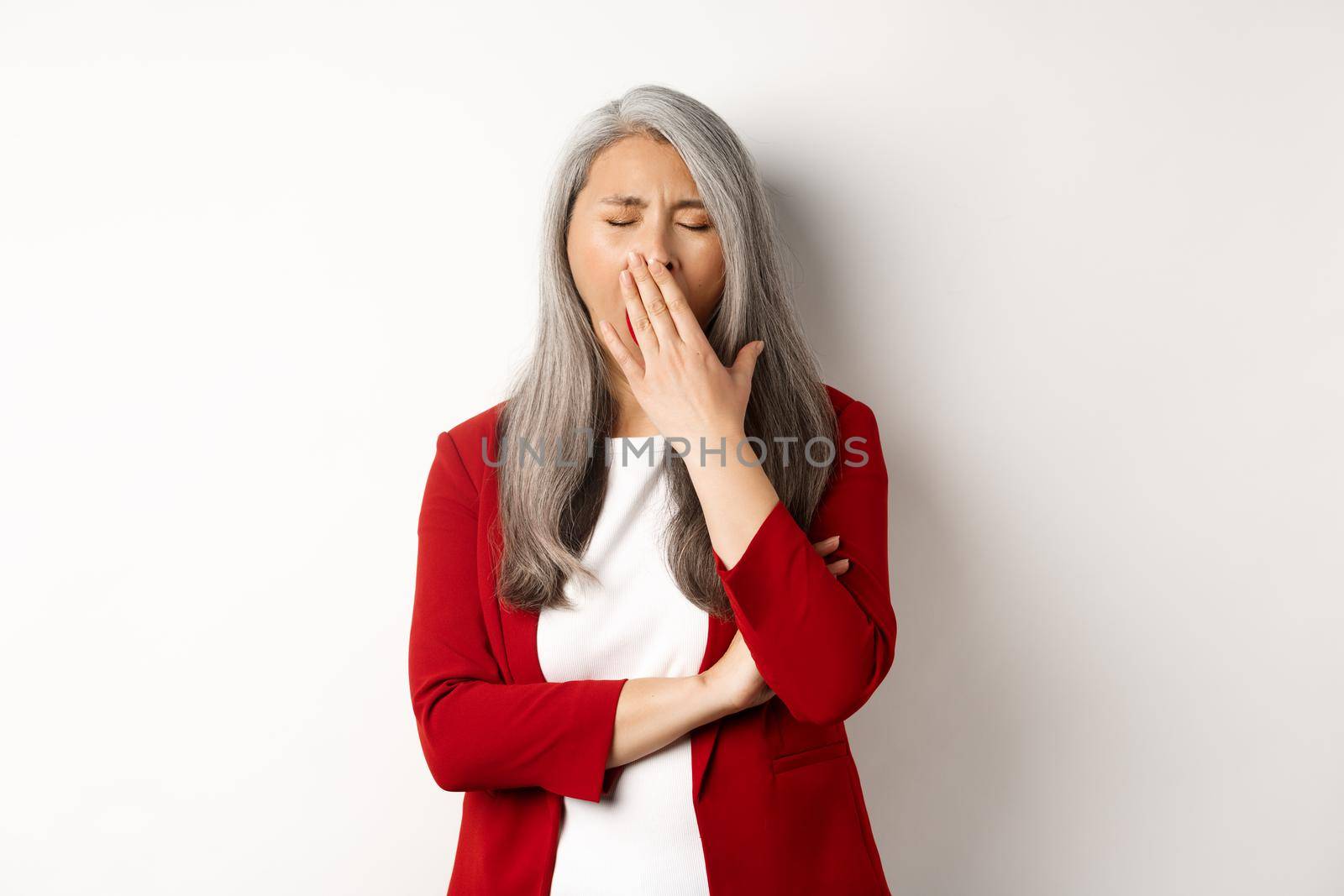 Tired or bored asian businesswoman yawning, covering mouth with hand, standing exhausted over white background by Benzoix