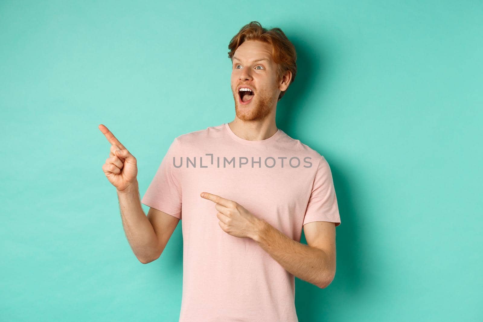 Amazed redhead man in t-shirt checking out promo, gasping in awe and pointing fingers at upper left corner, standing over mint background by Benzoix