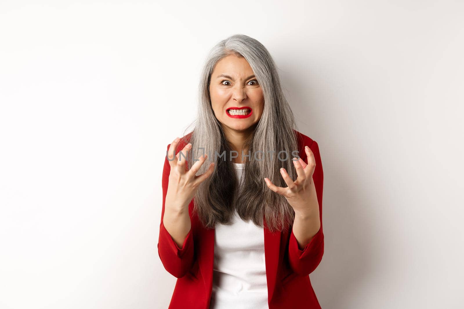Angry and pissed-off asian senior woman clench fists, grimacing and staring outraged, express hatred and annoyance, standing against white background by Benzoix