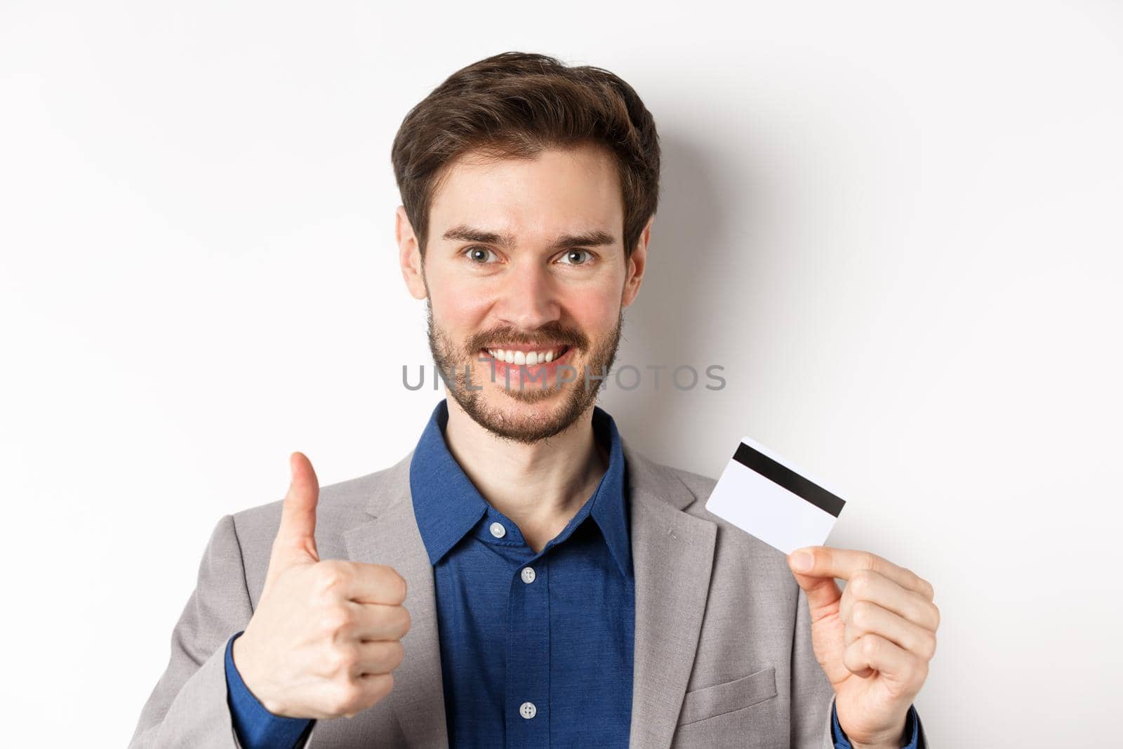 Shopping. Handsome satisfied client showing thumbs up and plastic credit card, smiling pleased, white background by Benzoix