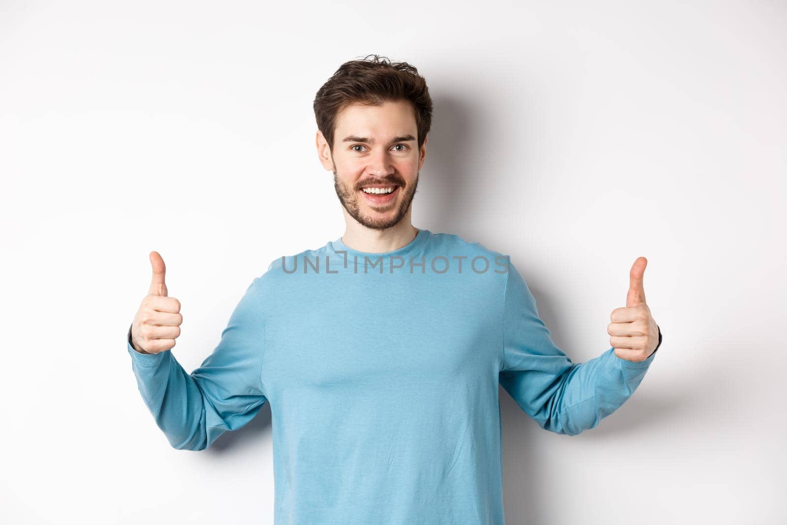 Cheerful young man showing thumbs up and smiling, recommend good product, praising choice, standing on white background.