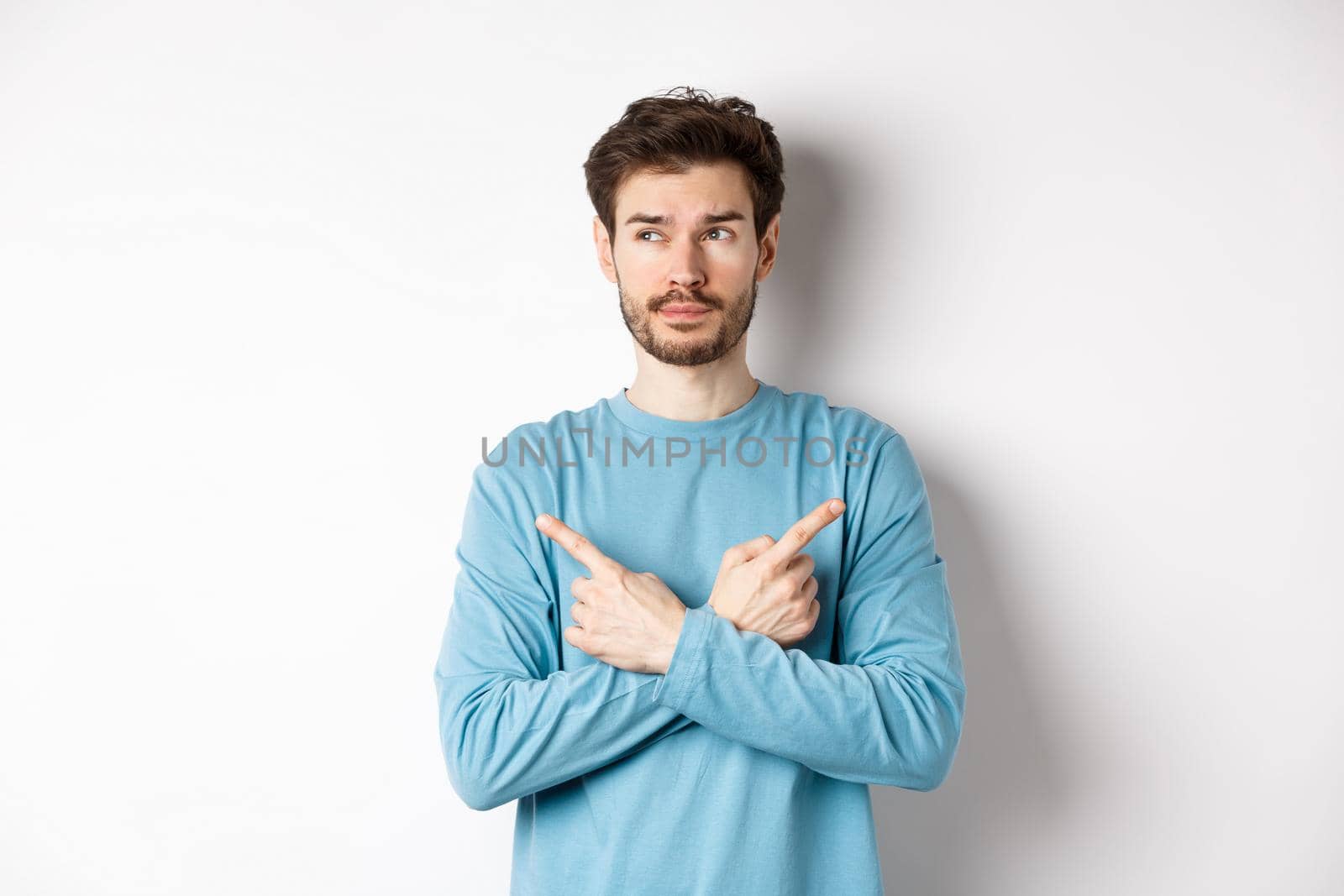 Indecisive man making choice, pointing fingers sideways at two variants and deciding, looking pensive at left side, standing on white background.