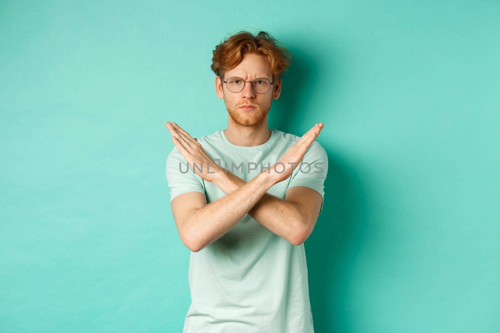 Serious and confident redhead man in t-shirt and glasses saying no, showing cross gesture to stop you, refucing or declining something, standing over turquoise background.