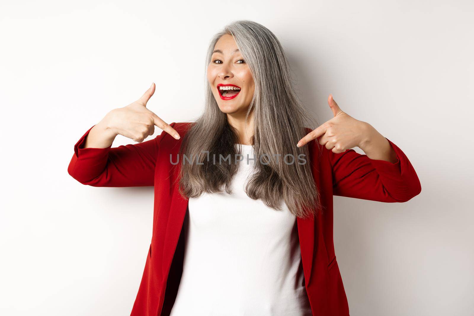 Ambitious senior woman pointing fingers at center, showing logo and smiling, standing over white background by Benzoix