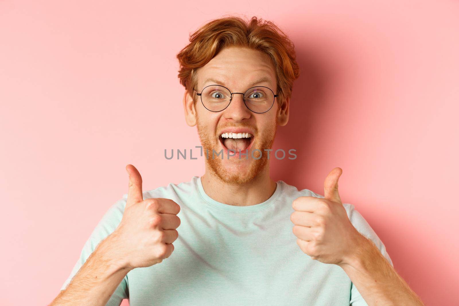 Face of happy redhead man in glasses and t-shirt, showing thumbs-up and looking excited, approve and praise cool promotion, standing over pink background.