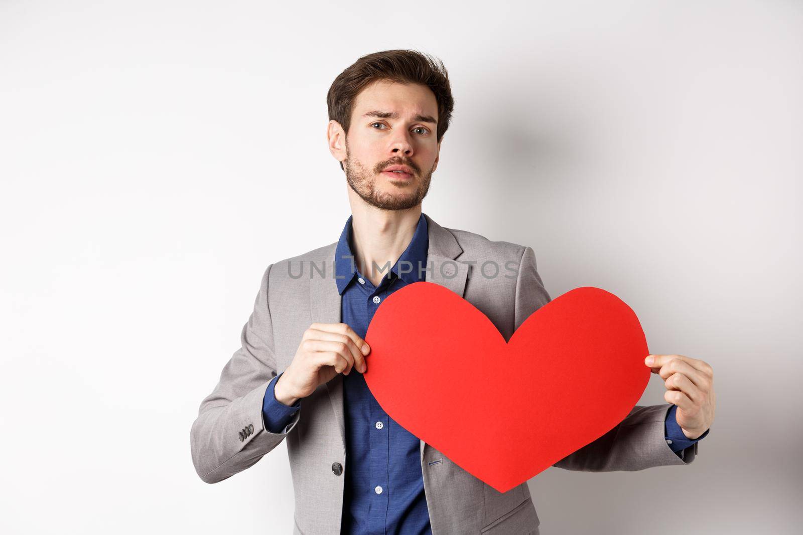 Passionate handsome man showing heart pounding gesture with red valentines cutout, standing in suit and searching for love, standing over white background by Benzoix