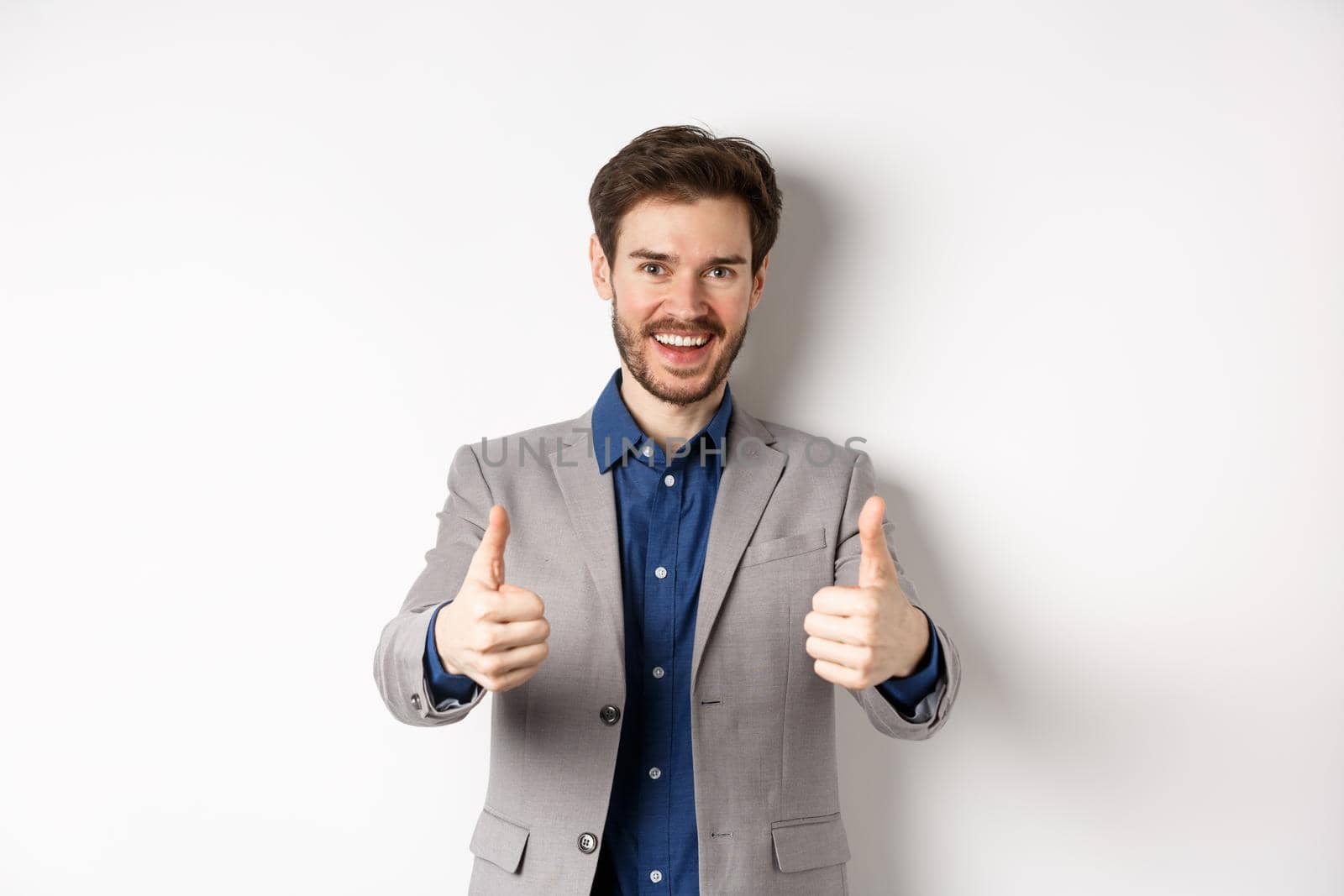 Cheerful and positive young man in business suit showing thumbs up and smiling, like something good, praise good job, standing happy on white background.