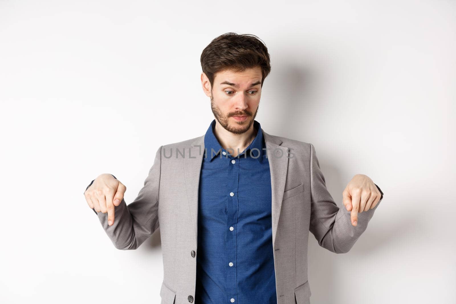 Surprised businessman looking and pointing down with raised eyebrows, see something curious, standing on white background in suit.
