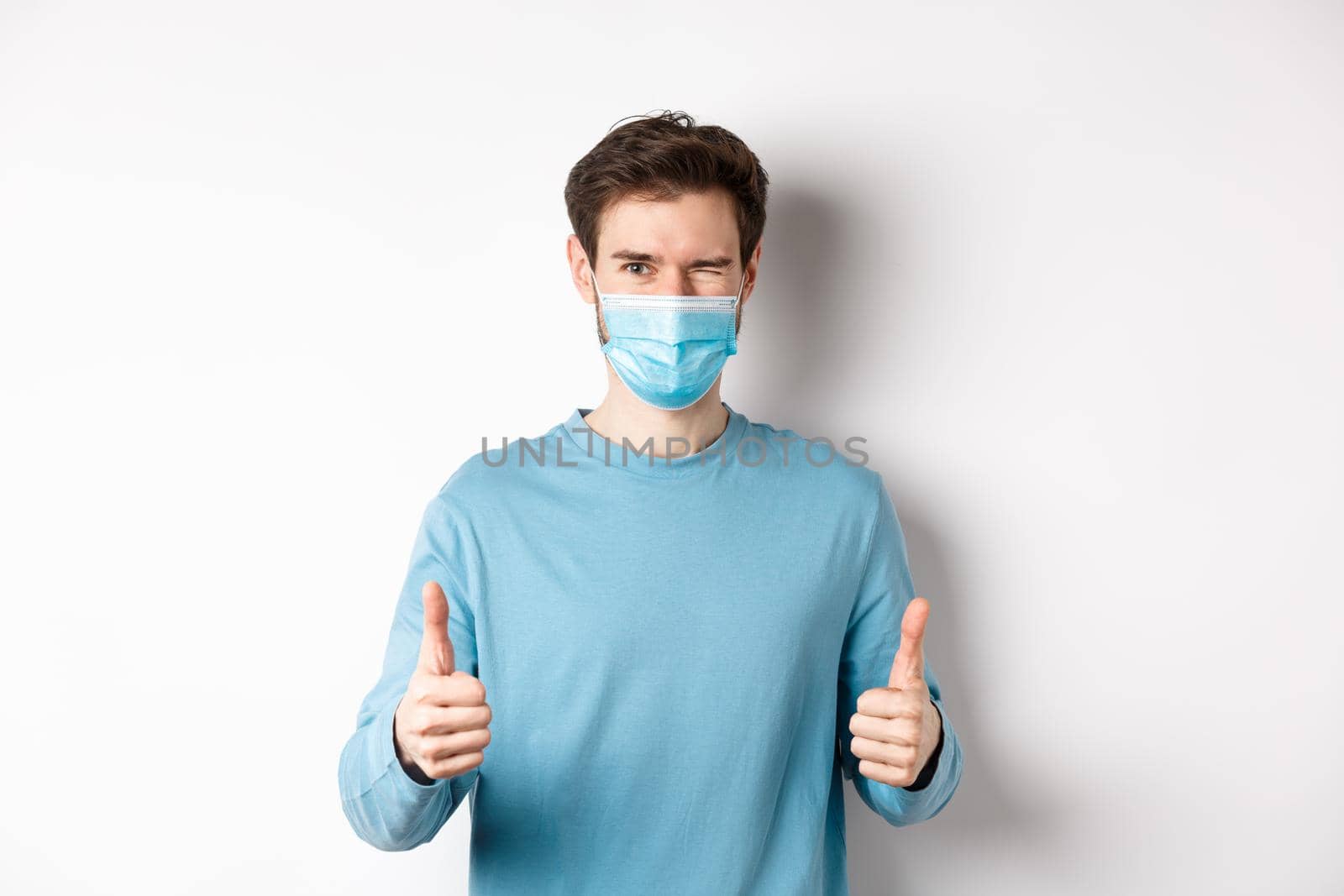 Covid-19, pandemic and social distancing concept. Happy young man in medical mask winking, showing thumbs up in approval, recommending product, white background.