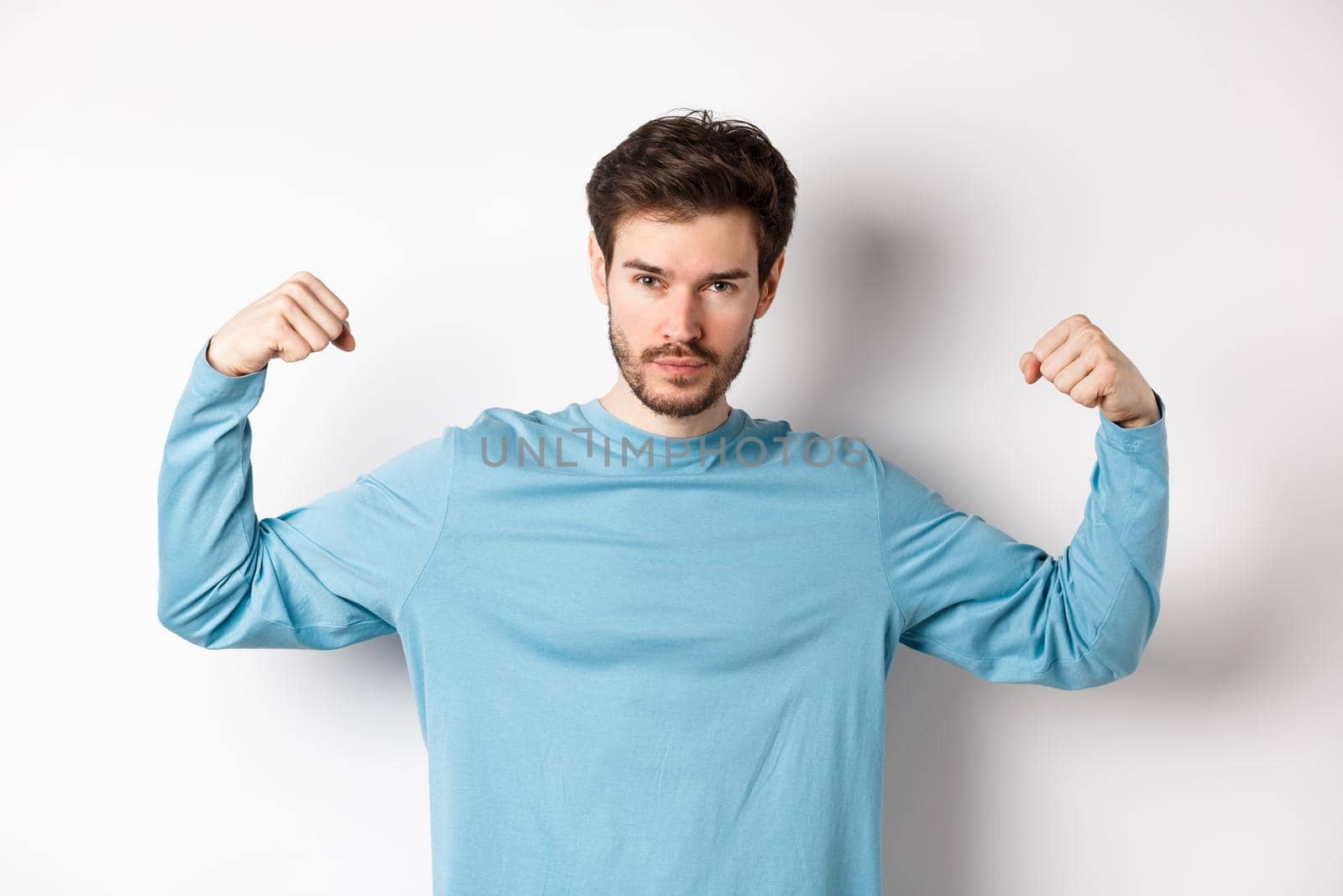 Confident and strong macho man flexing biceps, showing strength in muscles after gym workout, standing over white background.