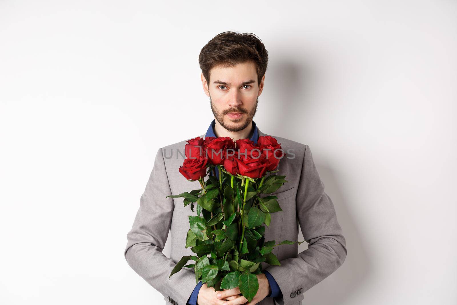 Valentines day and love concept. Confident and handsome man in suit holding bouquet of roses, looking at camera, standing over white background.