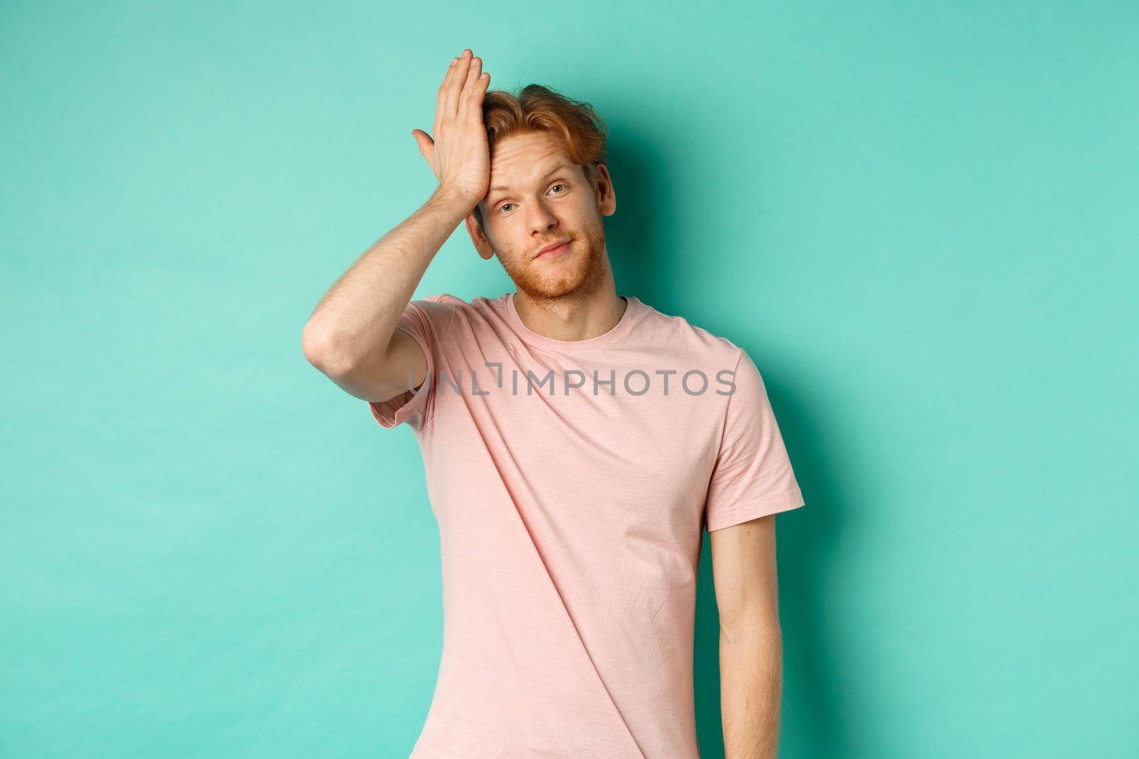 Annoyed and bothered redhead male model showing facepalm gesture, standing over mint background.