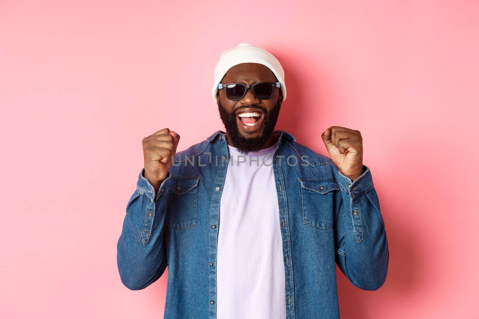 Satisfied african-american man in sunglasses saying yes, triumphing and making fist pumps in rejoice, celebrating achievement, standing like winner over pink background by Benzoix