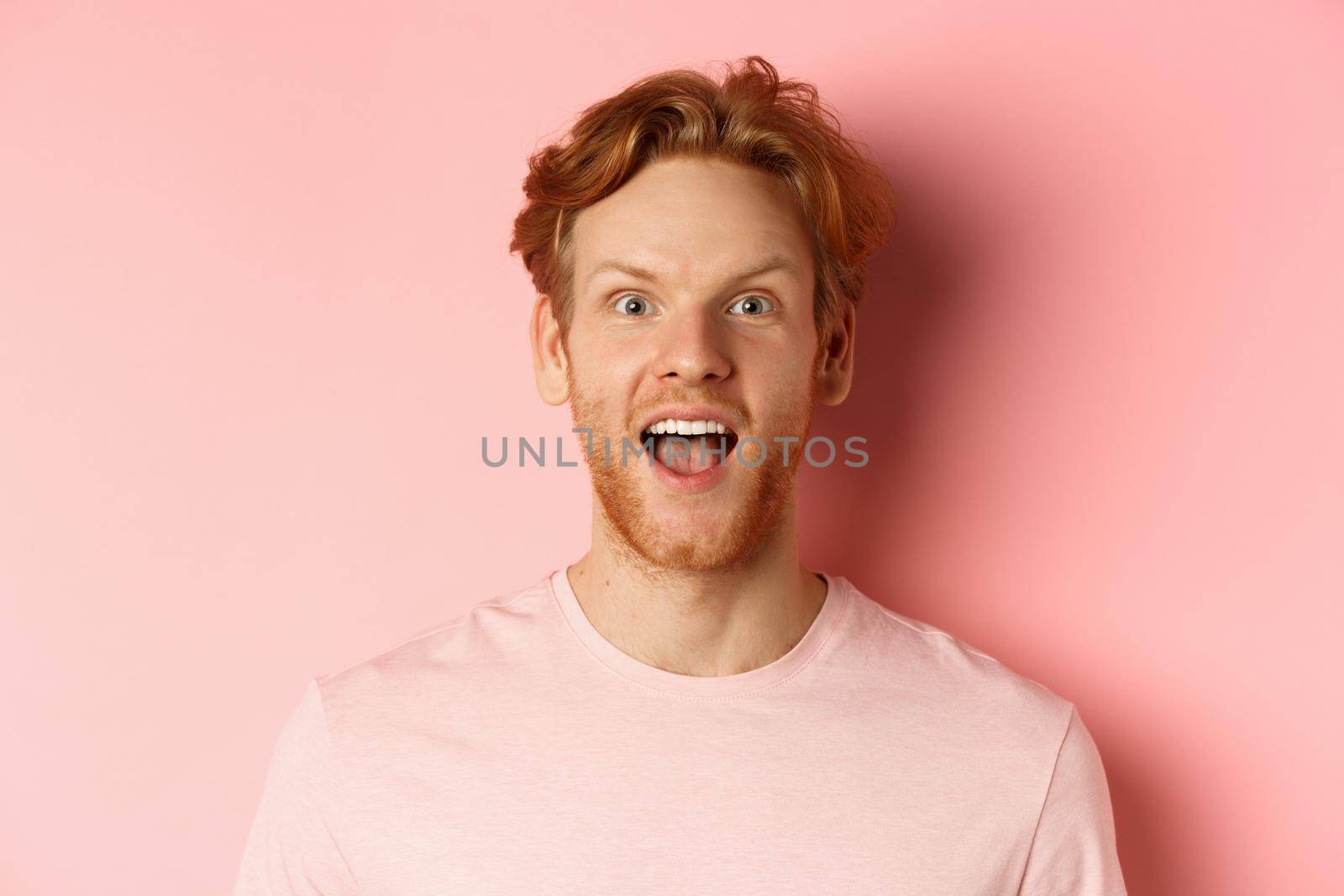 Headshot of amazed redhead male model in t-shirt open mouth, looking fascinated and happy at camera, standing over pink background by Benzoix