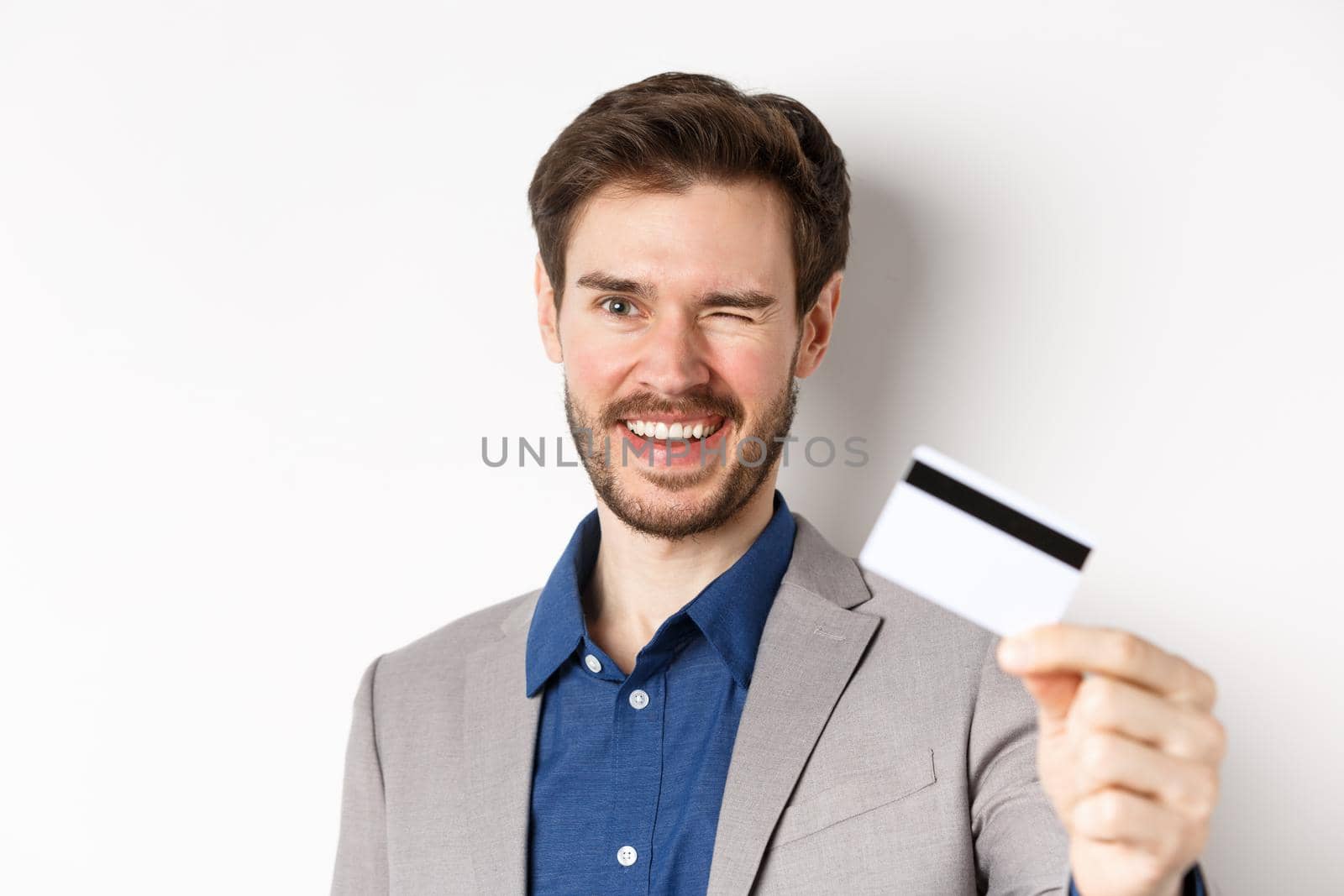Shopping. Cheerful bearded guy in suit winking and smiling, showing plastic credit card at camera, white background.