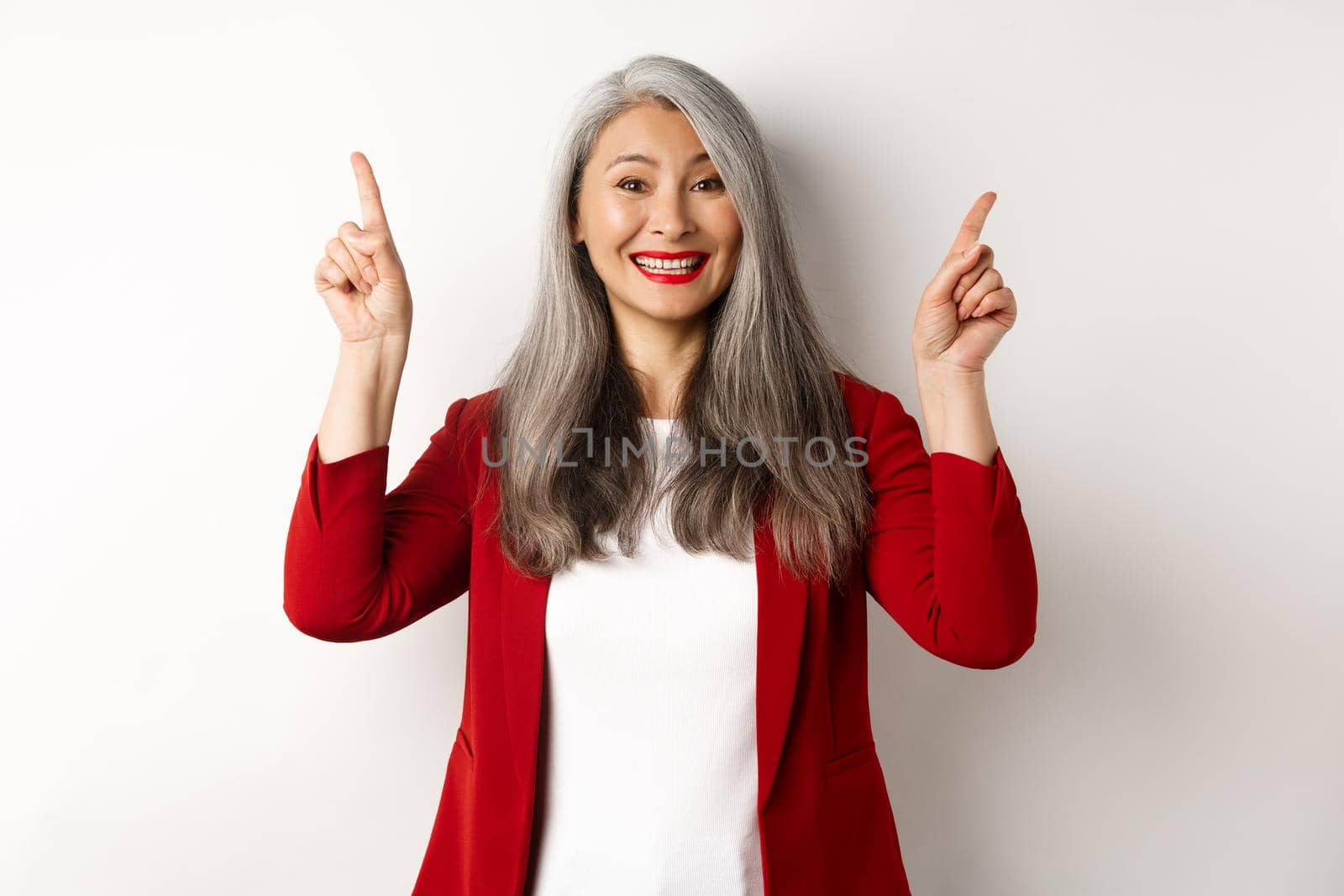 Happy mature woman in red blazer and makeup, smiling and showing advertisement on top, pointing fingers up at logo, white background.