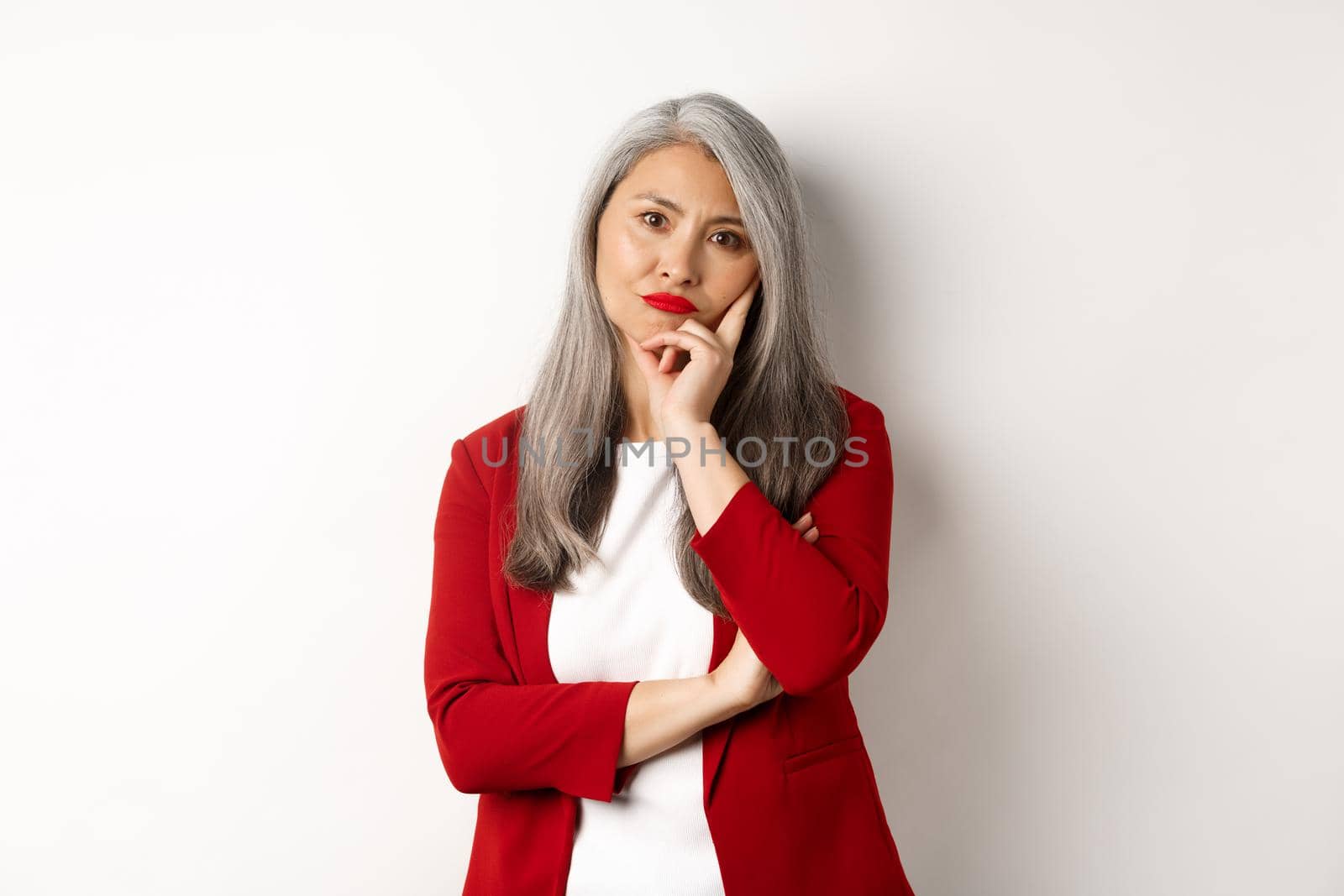 Annoyed and bothered asian businesswoman in red blazer, pouting and looking irritated at camera, standing over white background.