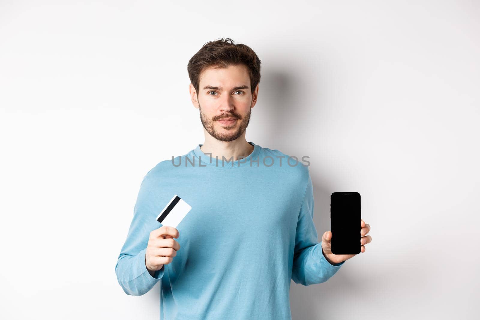Handsome caucasian man showing empty smartphone screen and plastic credit card, standing over white background by Benzoix