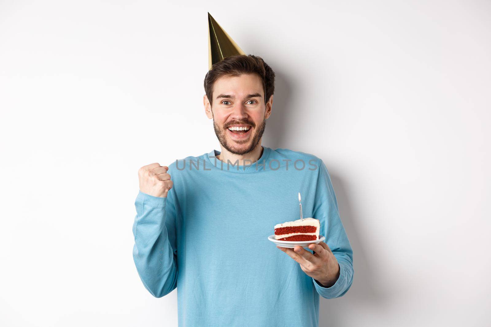 Celebration and holidays concept. Cheerful young man celebrating birthday in party hat, saying yes and fist pump in joy, holding bday cake, white background.