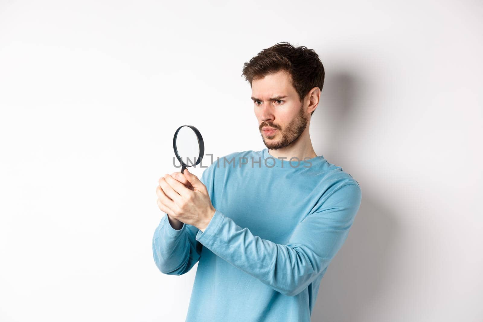 Serious-looking man looking through magnifying glass, investigating something, found interesting promo, standing on white background.