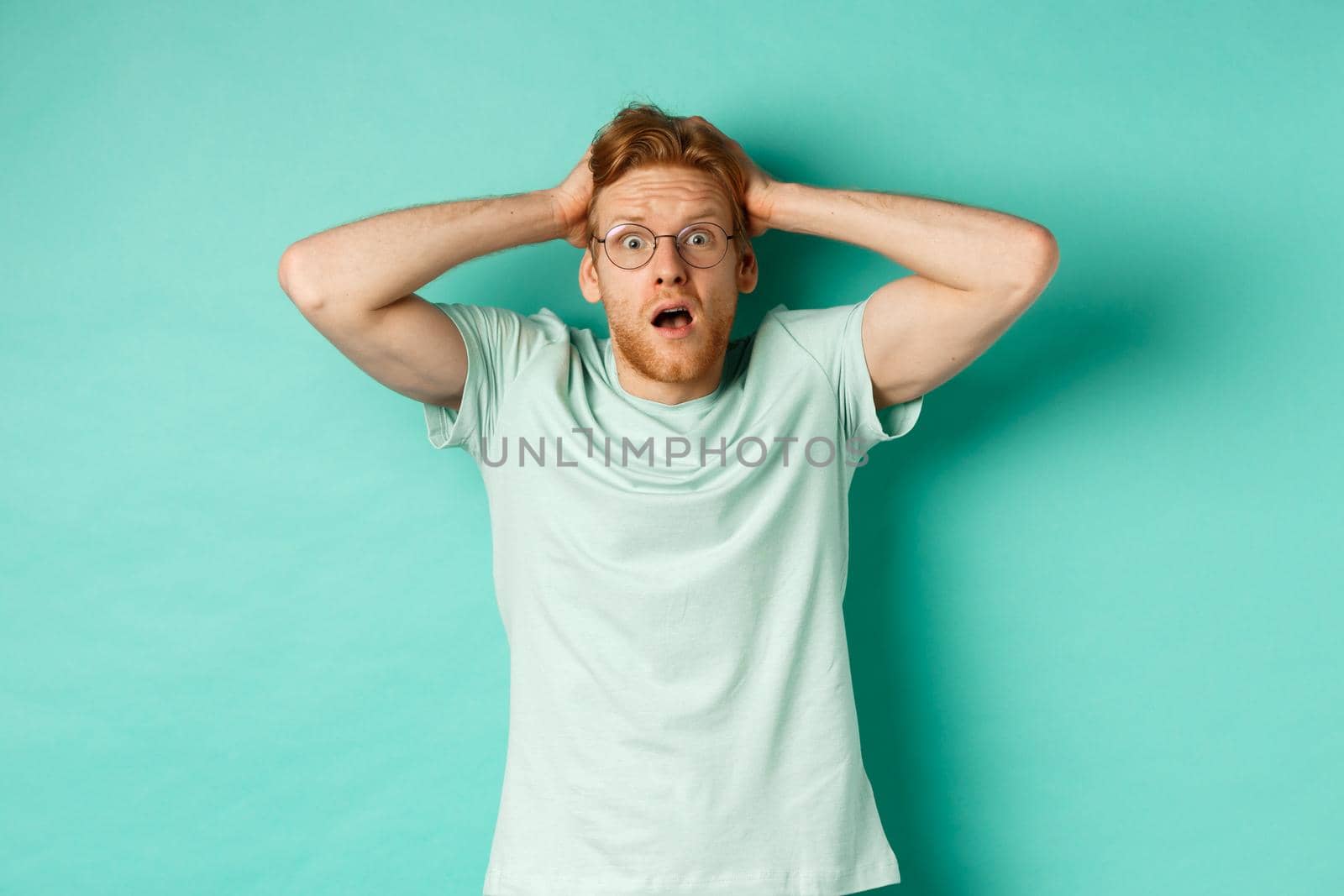 Young redhead man with beard, wearing glasses and t-shirt, holding hands on head and staring in panic, standing alarmed and anxious against turquoise background by Benzoix