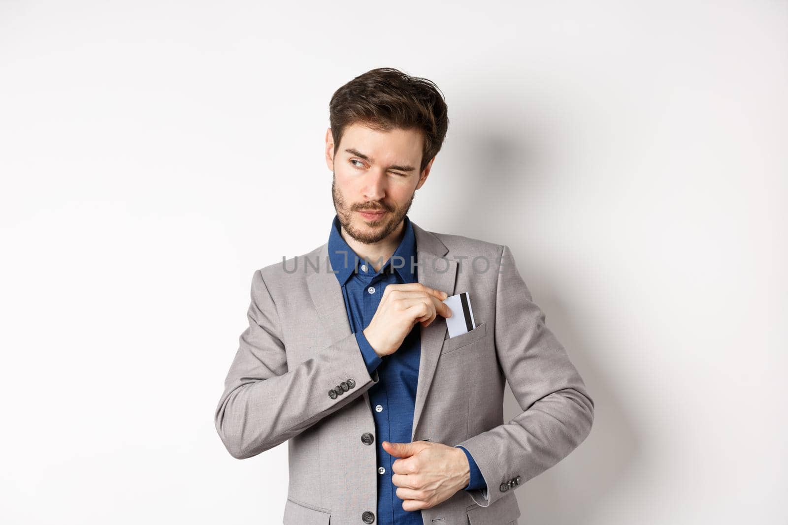 Confident and successful businessman winking aside while place credit card in suit pocket, standing on white background.