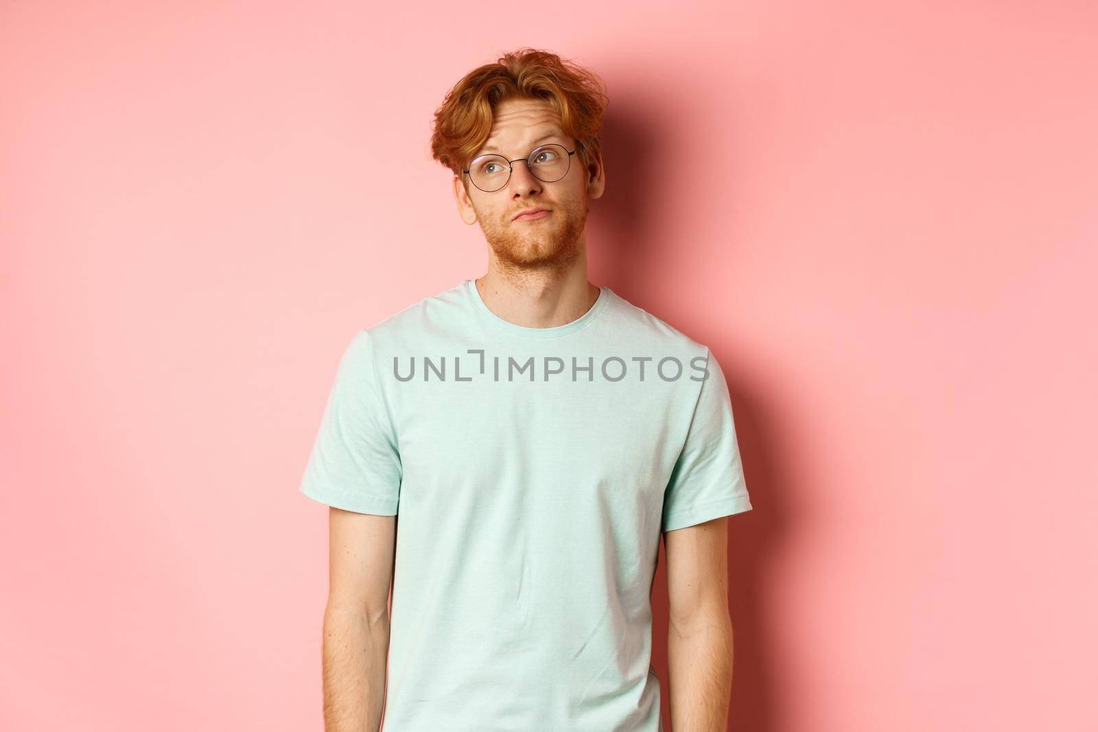 Image of young redhead man with beard, wearing t-shirt and glasses, looking left with bored unbothered face, standing over pink background by Benzoix