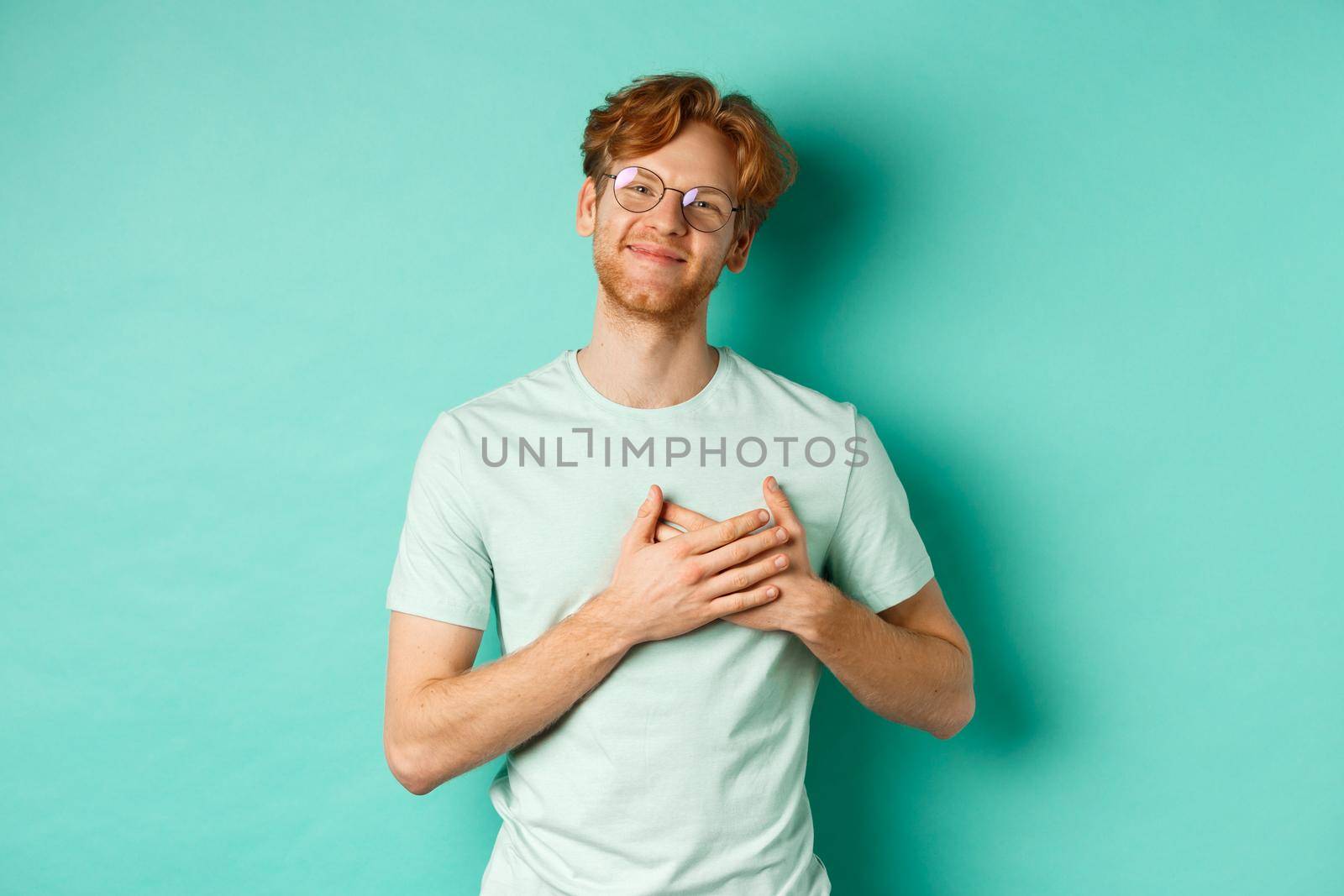 Handsome young man with red hair and glasses, holding hands on heart and smiling, saying thank you, feeling grateful and touched, standing over turquoise background by Benzoix