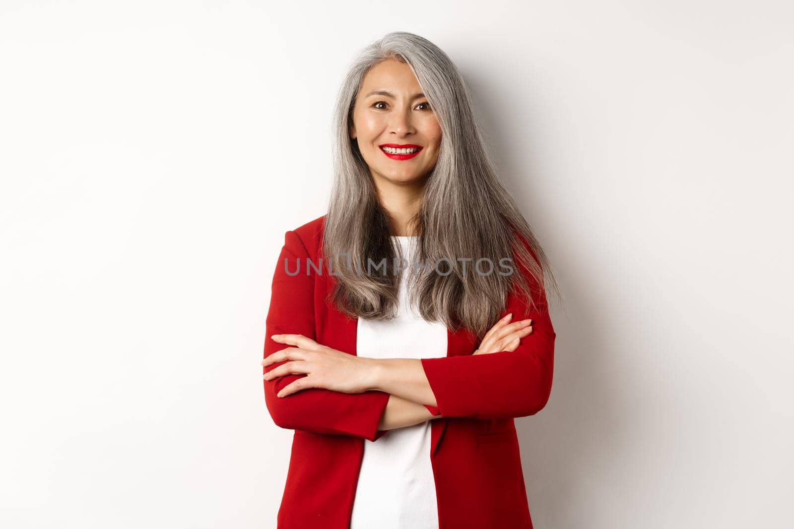 Business people. Smiling asian senior woman in red blazer, cross arms on chest and looking professional, standing over white background by Benzoix