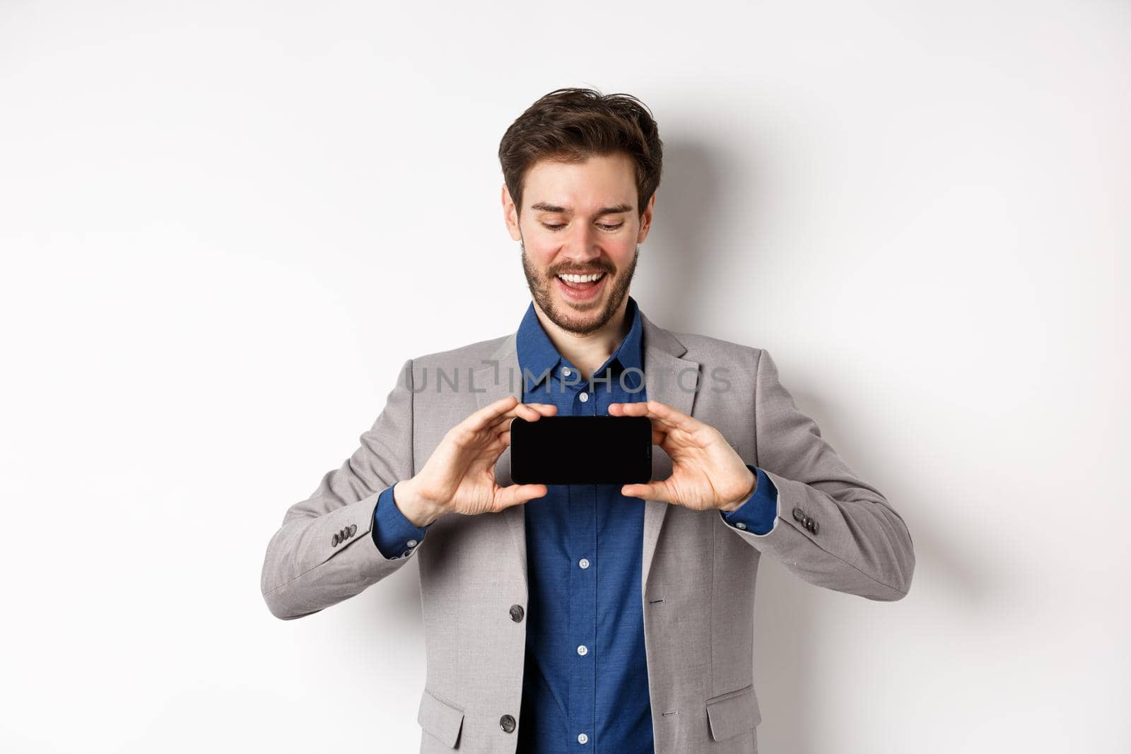 E-commerce and online shopping concept. Happy young man in suit laughing, showing empty smartphone screen horizontally, wite background by Benzoix