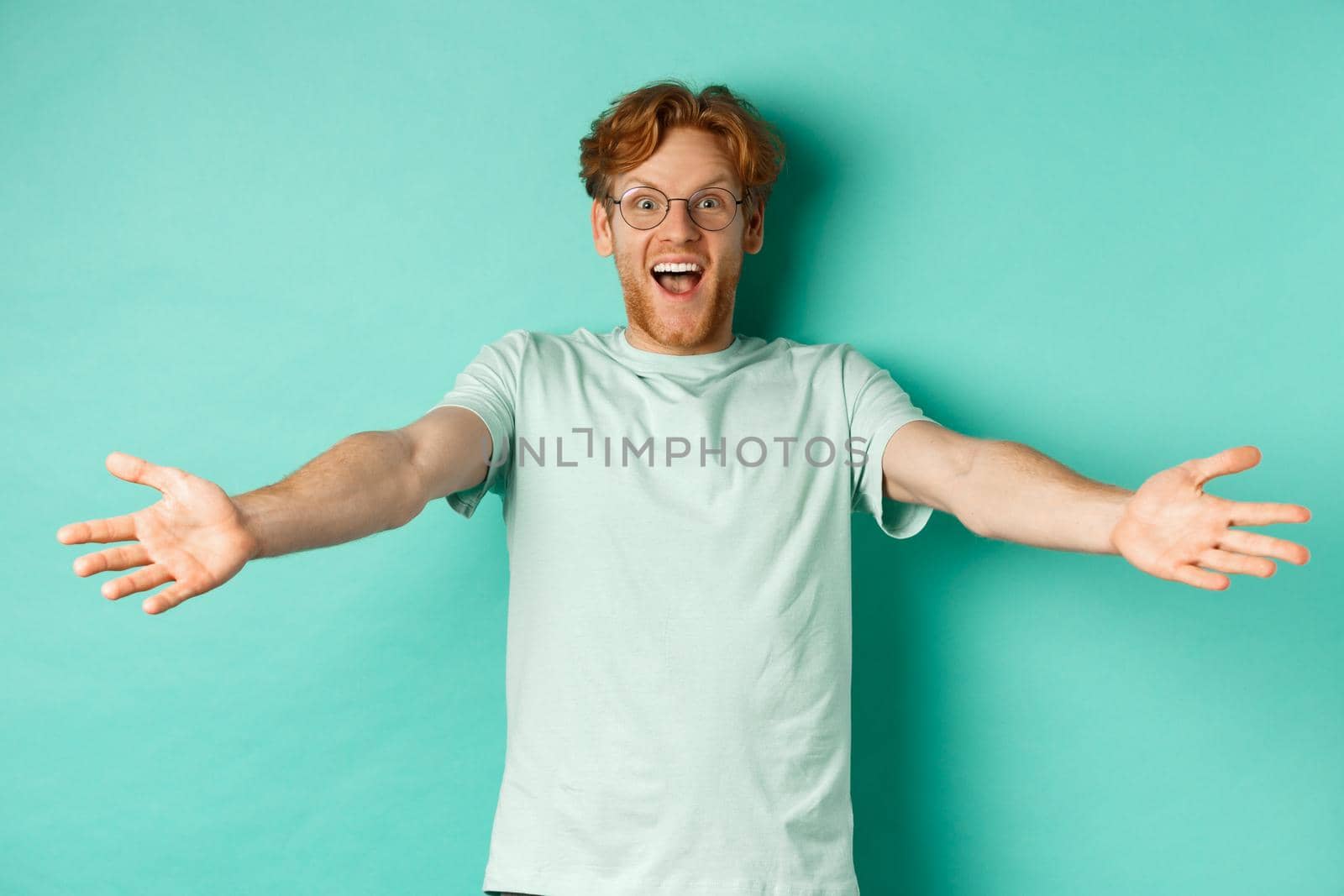 Excited young redhead guy in glasses strethced out hands in warm welcome, invite you and smile friendly at camera, standing happy over turquoise background.