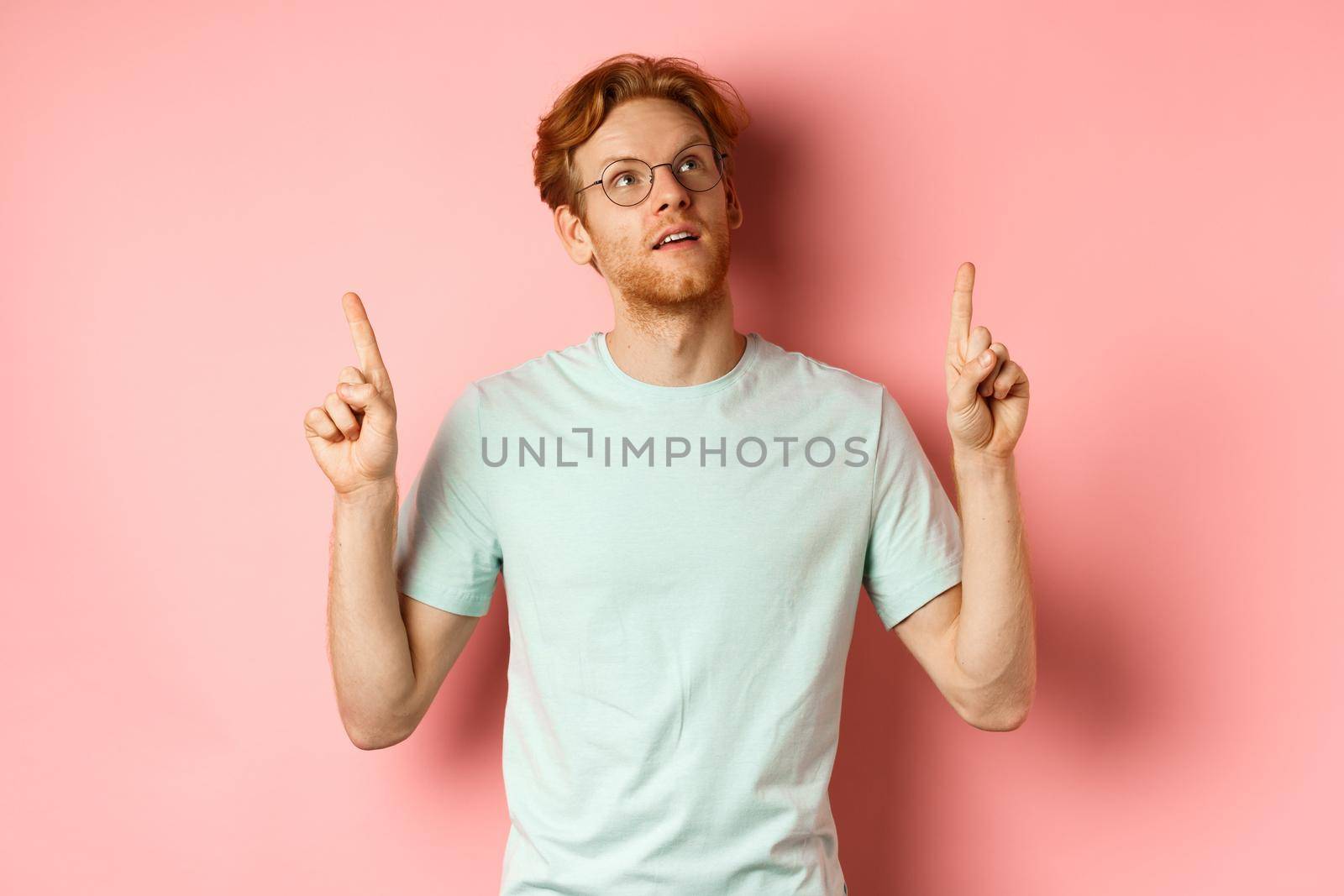 Dreamy young man with red hair and beard pointing fingers up, looking pensive at top, standing against pink background by Benzoix