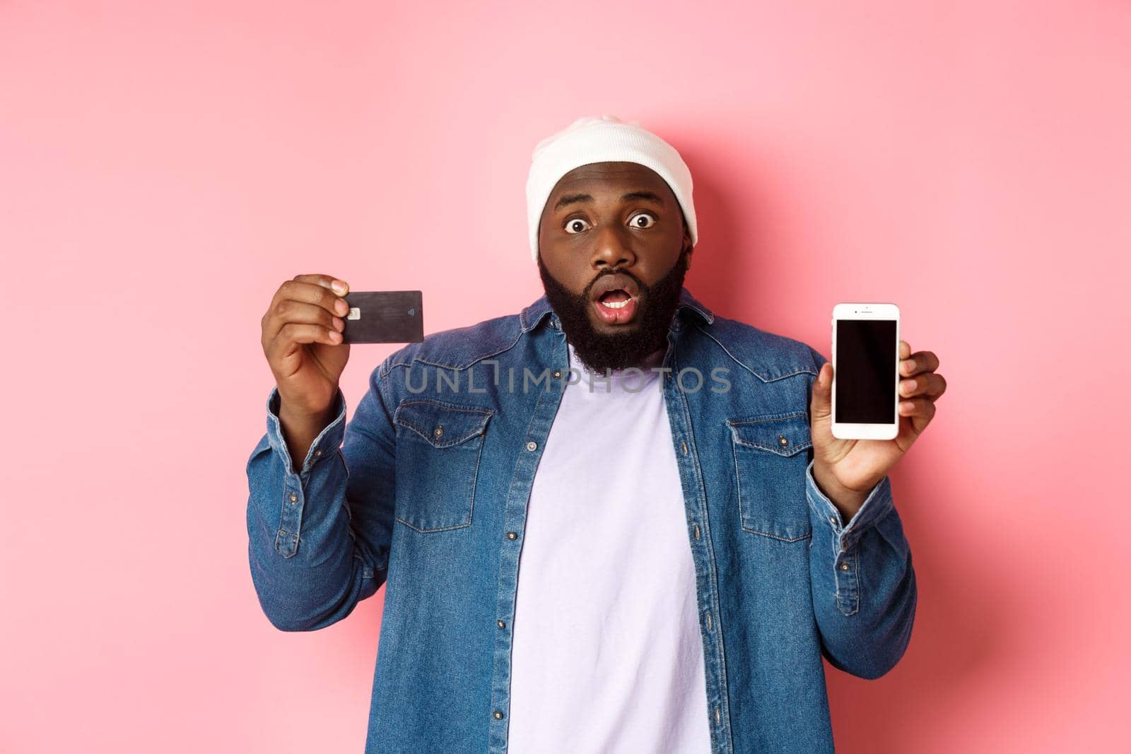 Online shopping. Shocked and concerned Black man staring at camera, showing mobile phone screen and credit card, standing over pink background by Benzoix