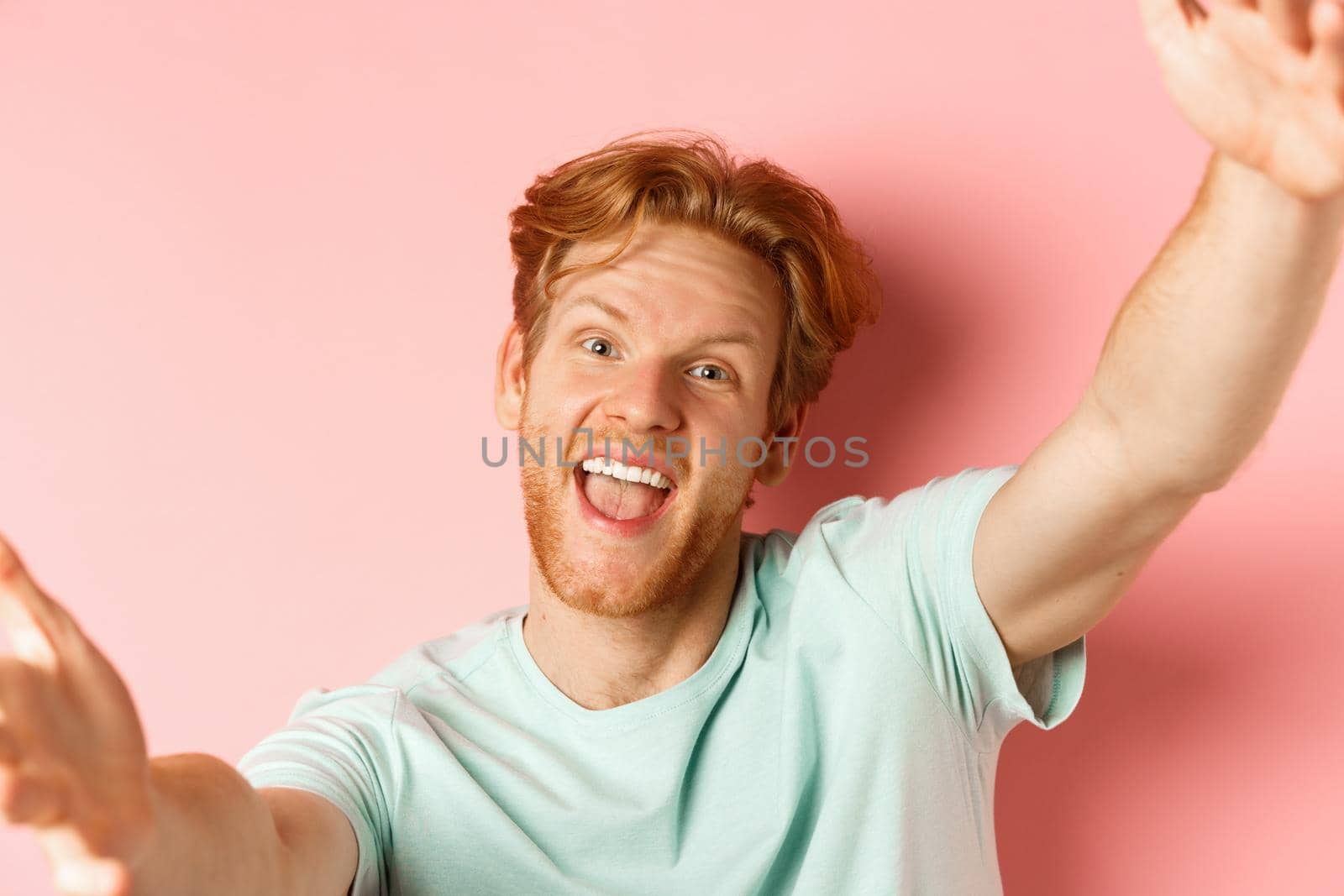 Funny handsome redhead man taking selfie, stretch out hands to hold camera and smiling happy, look from smartphone, standing over pink background.